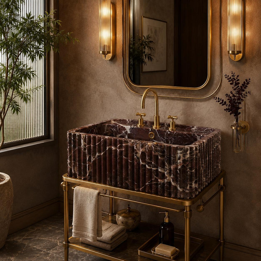 A luxury bathroom vanity featuring a rectangular marble sink with dark reddish-brown and white veining, mounted on a polished gold brass stand. The setup includes a matching gold faucet, gold wall sconces, a large decorative mirror, and a small shelf holding folded towels and toiletries, all set against a textured warm brown wall.