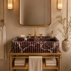 A luxury bathroom vanity setup featuring a rectangular ribbed marble sink in a deep burgundy and gold pattern, mounted on a gold metal console table. The scene is styled with a matching gold-framed mirror, warm wall sconces, beige towels, and a decorative vase with dried branches.