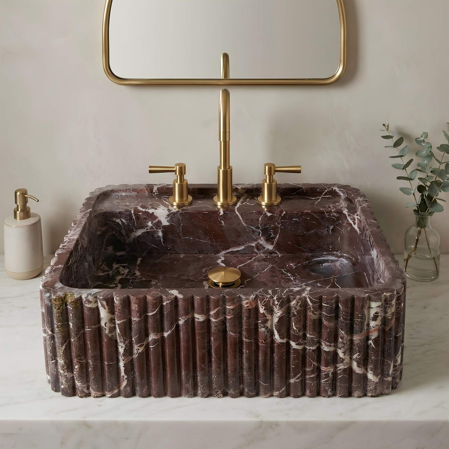 A rectangular vessel sink made of dark brown marble with white veining, featuring a fluted front edge, set on a white marble countertop with a matching gold faucet, gold pop-up drain, and a gold-framed mirror above.