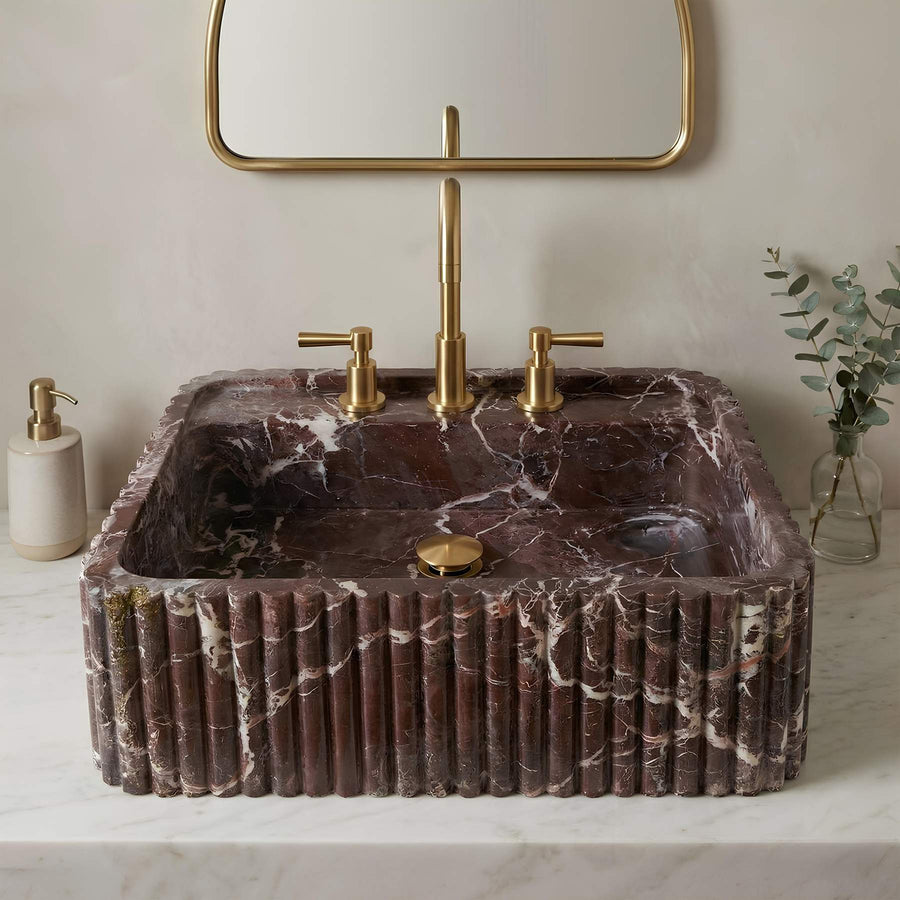 A rectangular vessel sink made of dark brown marble with white veining, featuring a fluted front edge, set on a white marble countertop with a matching gold faucet, gold pop-up drain, and a gold-framed mirror above.