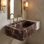 A modern wall-mounted sink with a dark reddish-brown fluted marble basin and brushed gold faucet and trim, shown installed against a beige plaster wall with a matching mirror and sconces.