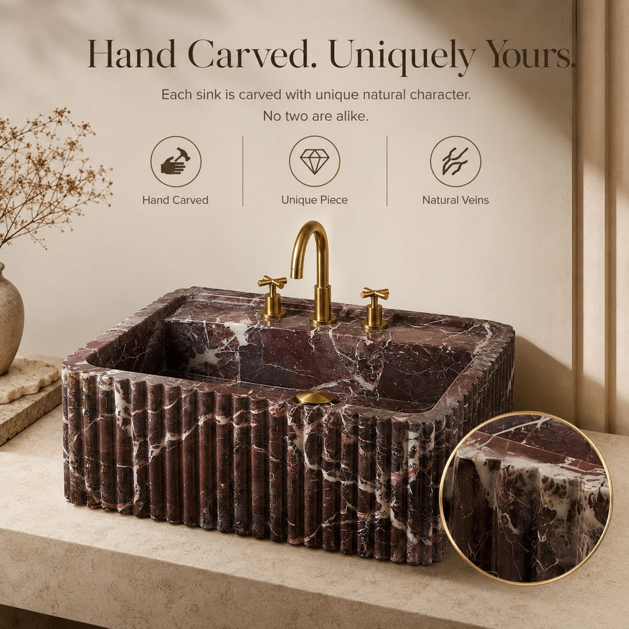 A rectangular vessel sink made of dark reddish-brown marble with white veining, featuring a fluted front edge and a curved gold faucet. The sink is shown on a beige countertop with a matching marble backsplash, a decorative vase with dried branches, and marketing text above with a circular inset showing a close-up of the marble texture.