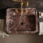 A rectangular dark red-brown marble sink with white veining, featuring a matching brass faucet and cross handles, set into a dark marble countertop with folded white towels on either side.