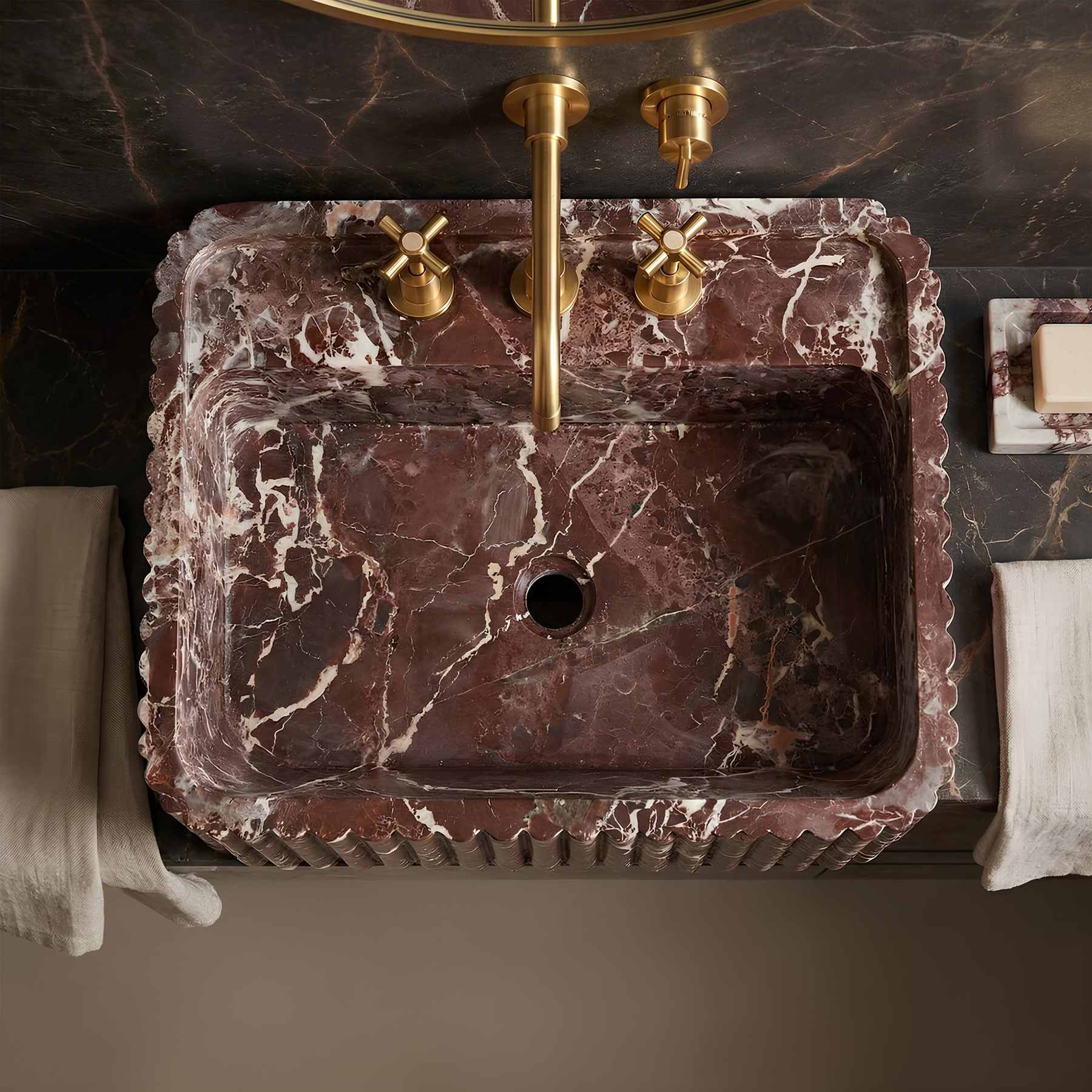 A rectangular dark red-brown marble sink with white veining, featuring a matching brass faucet and cross handles, set into a dark marble countertop with folded white towels on either side.